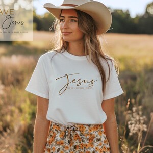 Könnte beinhalten: Eine Frau trägt ein weißes T-Shirt mit dem Wort "Jesus" in brauner Schrift. Der Text "The Way, The Truth, The Life" steht unter dem Wort Jesus. Sie trägt einen Cowboyhut und einen Blumenrock. Das Bild hat einen warmen, natürlichen Hintergrund.