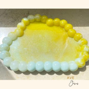 Può includere: Un vassoio decorativo a forma di cuore, con una sfumatura di colori dal bianco al giallo. Il bordo del vassoio è composto da elementi arrotondati a forma di perla. Il testo "AVE Jesus" è scritto in corsivo in basso.