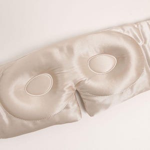 May include: A champagne-coloured sleep mask with a smooth, lustrous finish. The mask features two contoured, padded eye sections with stitched oval outlines, designed to sit over the eyes, connected by a wide, flexible band of matching fabric.