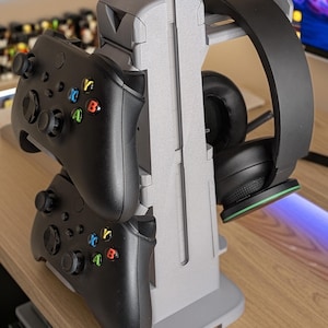 May include: A gray 3D-printed gaming accessory stand holding two black game controllers and a black headset. The stand is designed to organize gaming equipment and features a sturdy base and multiple slots.