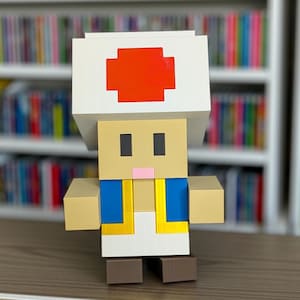 May include: A 3D-printed figure of the character Toad from the Super Mario video game series. The figure features a white mushroom cap with a red cross, a tan face, and a blue vest with yellow trim. The figure is standing on a brown surface.