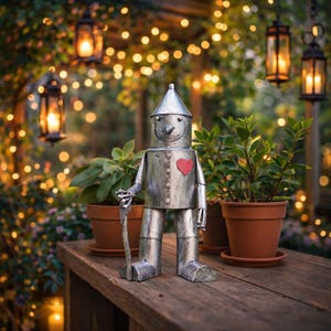 Może przedstawiać: Metalowa rzeźba Blaszaneho Człowieka z czerwonym sercem i laską. Srebrna figura stoi na drewnianej powierzchni, otoczona roślinami doniczkowymi i wiszącymi lampionami.