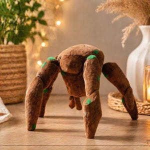 May include: A brown plush spider toy with six legs and a rounded body. The legs and body have a textured appearance, with green accents on the leg joints. The toy is designed to stand on its legs.