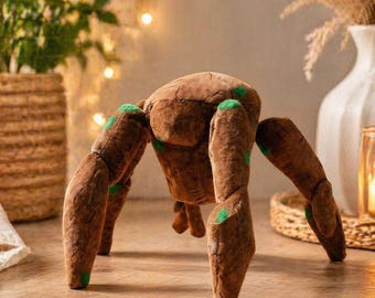 May include: A brown plush spider toy with six legs and a rounded body. The legs and body have a textured appearance, with green accents on the leg joints. The toy is designed to stand on its legs.