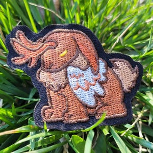 May include: Embroidered patch depicting a cartoon mammoth with brown fur, orange tusks, and a white chest. The patch has a black border, set against green grass.
