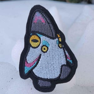 May include: Embroidered patch of a stylized cat face with a light blue body, yellow eyes, and pink and blue accents. The patch has a black border, set against a snowy background.