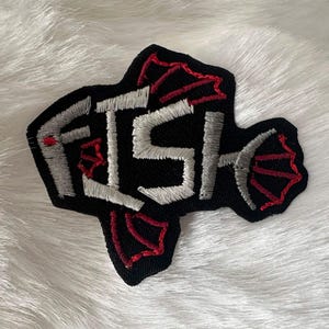 May include: A black embroidered patch in the shape of a fish, featuring the word FISH stitched in light gray thread. The design includes red thread accents outlining the fins and tail, creating a bold contrast against the black fabric background.