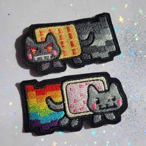 May include: Two embroidered patches featuring pixelated cat designs. One cat has a yellow and orange background, the other has a rainbow background. Both cats have gray bodies, pink cheeks, and angry red eyes.