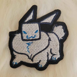 May include: Embroidered patch featuring a stylized white rabbit with a square head and blue accents. The patch has a black border and is likely for clothing or accessories.