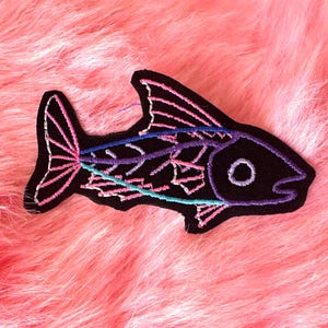 May include: A black embroidered fish patch featuring a skeletal design outlined in vibrant neon pink, purple, and teal thread. The fish shape is clearly defined with a prominent dorsal fin, a tail fin, and a circular eye, all stitched onto a black fabric base.
