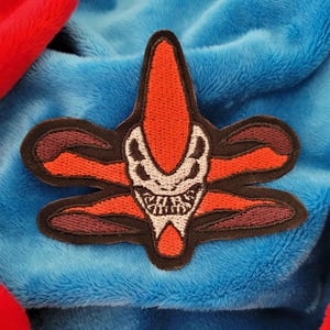 May include: Embroidered patch featuring a stylized, orange and black alien-like creature with a white skull-like face and sharp teeth. The patch is set against a blue and red plush background.