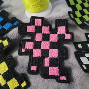 May include: Embroidered patches in various colors, including pink, blue, yellow, green, and white, with a black checkered design. The patches are arranged on a gray surface, with a spool of yellow thread in the background. These patches are suitable for clothing or accessories.