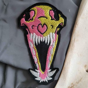 May include: Embroidered patch depicting a stylized skull with a heart detail. The skull is pink and yellow, with white teeth and a black border. The patch is set against a grey background.