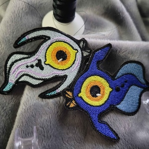 May include: Two embroidered patches with a bird-like design. One is light grey with purple and teal accents, and the other is blue with yellow and orange details. Both have a black border and a large eye in the centre.