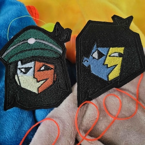 May include: Two embroidered patches with character designs. One features a face with a green hat, white, and orange sections. The other patch has a blue and yellow face. Both are on black backgrounds with orange thread.