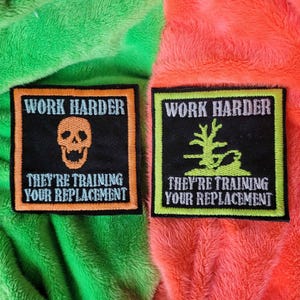 May include: Two embroidered patches. One features an orange skull, the other a green tree, both on black backgrounds. Text reads "WORK HARDER" and "THEY'RE TRAINING YOUR REPLACEMENT". Contrasting borders frame the text and images.