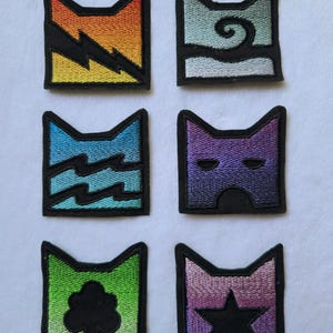 K&ouml;nnte beinhalten: Sechs quadratische Stickaufn&auml;her mit schwarzem Rand, die jeweils eine Katzenohren-Silhouette zeigen. Die Motive umfassen einen Blitz auf Orange, eine Spirale auf Hellblau, Wellen auf Blau, eine Maske auf Violett, einen Baum auf Gr&uuml;n und einen Stern auf Pink-Violett.