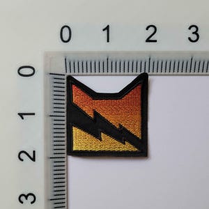 K&ouml;nnte beinhalten: Ein quadratischer gestickter Aufn&auml;her mit einem schwarzen Blitz auf einem Farbverlauf, der von Orange oben zu Gelb unten &uuml;bergeht. Der Aufn&auml;her ist an einem Lineal ausgerichtet, das eine Breite von etwa 3,8 cm anzeigt.