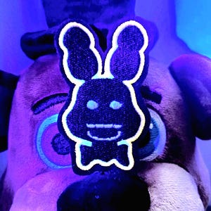 May include: A black and white bunny patch with a smiling face and bow tie is centered in front of a plush bear. The bunny patch has a white outline and is set against a purple and blue background.