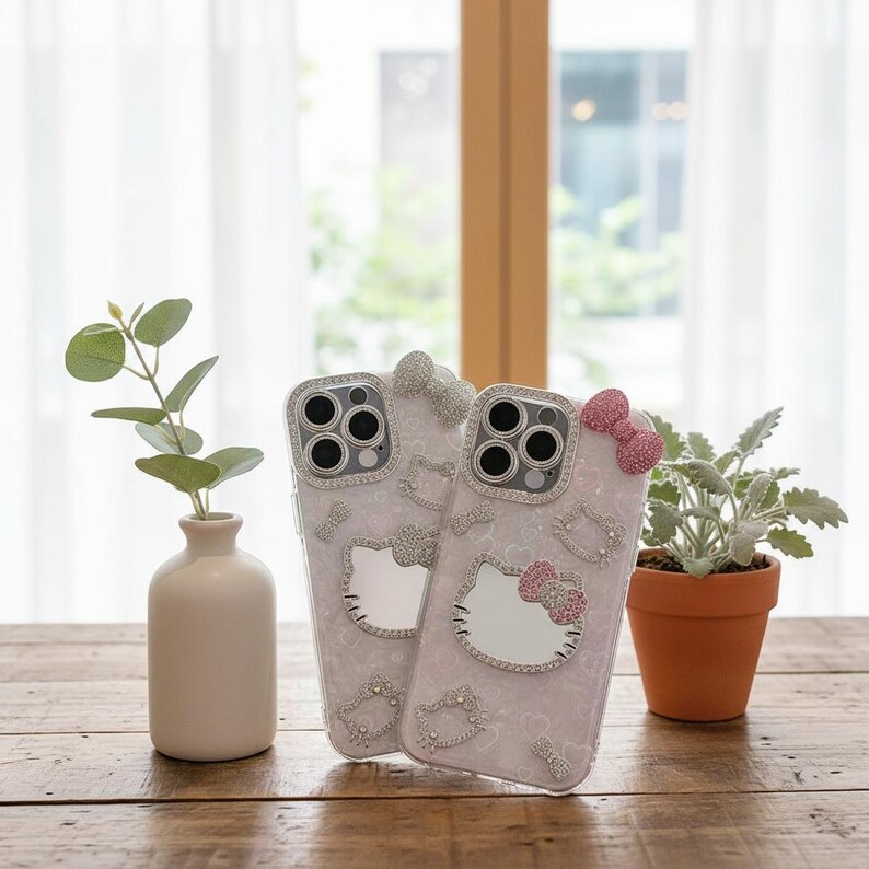 May include: Two clear phone cases with a Hello Kitty design. The cases feature a pink and white pattern, embellished with rhinestones and bows. The cases are on a wooden table, with a small vase and a potted plant in the background.