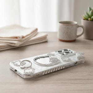 May include: A white and silver phone case with a decorative design. The case features a ring holder, a mirror, and several silver embellishments. The case is on a wooden surface, with a mug, plant, and folded cloth in the background.