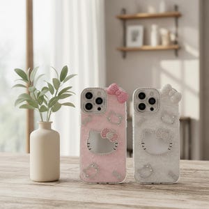 May include: Two phone cases with a Hello Kitty design. One case is light pink, the other is white. Both cases have a bow at the top and Hello Kitty's face in the center, embellished with rhinestones. The cases are on a wooden surface.