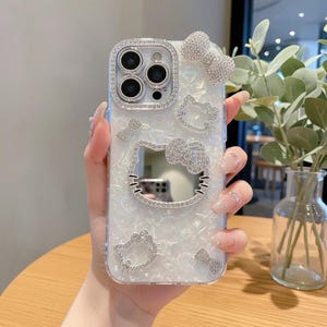 May include: A clear phone case with a pearlescent finish, adorned with sparkling rhinestones. The design includes a Hello Kitty outline, bows, and heart shapes. The camera lens is surrounded by a rhinestone border.