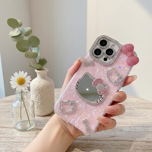 May include: A pink phone case with a Hello Kitty design. The case features a mirrored Hello Kitty face, rhinestone accents, and a pink bow. The case is designed to fit a smartphone.