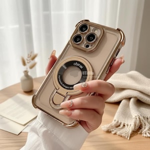 May include: A gold-colored phone case with a clear back and a circular ring holder. The case has a gold frame and a ring with the text "cbinei". The phone case is held in a person's hand.