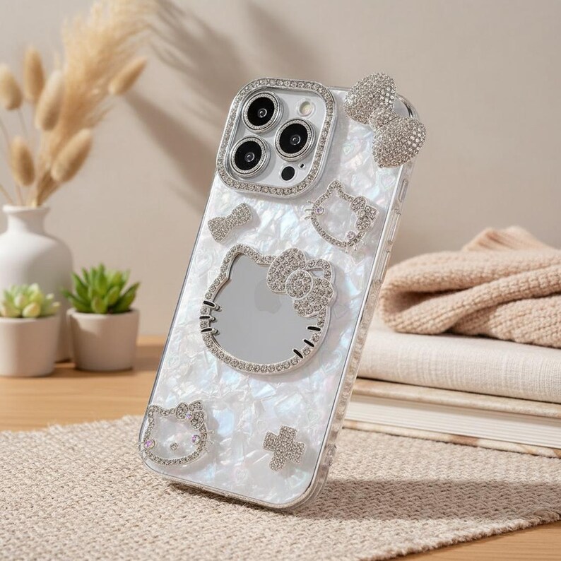 May include: Clear phone case with a pearlescent back featuring a Hello Kitty design. The case is adorned with rhinestones around the camera, Hello Kitty's face, bow, and other decorative elements. A bow-shaped embellishment is attached to the top.