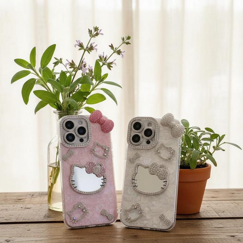 May include: Two phone cases with Hello Kitty designs. One is pink with a pink bow, the other is white with a silver bow. Both cases have a mirror Hello Kitty face, surrounded by rhinestones and other decorative elements.