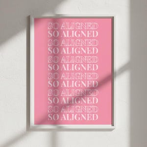 May include: A framed pink art print with the words "SO ALIGNED" repeated in white. The print is in a white frame and hangs on a white wall. The artwork has a minimalist design.