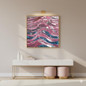 May include: Abstract art print in a gold frame. The artwork features wavy, layered shapes in shades of pink, blue, and silver, creating a shimmering effect. The print is displayed above a white console table with gold legs, and two pink ottomans.