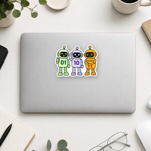 May include: A die-cut sticker featuring three cartoon robots standing side-by-side. The green robot on the left has the number 01, the purple robot in the centre has 10, and the orange robot on the right has 11 printed on their chests.