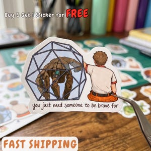 May include: A die-cut sticker featuring a brown, robotic creature inside a geometric, blue-lined cage, with a person in a white shirt and orange pants reaching toward it. The text reads, "you just need someone to be brave for".