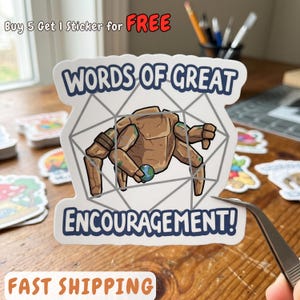 May include: A die-cut sticker featuring a brown, multi-limbed rock creature holding a small globe. The design is framed by a gray geometric outline and includes the dark blue text "WORDS OF GREAT ENCOURAGEMENT!" printed above and below the creature.