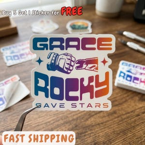May include: A die-cut sticker featuring a gradient color scheme from blue to orange. The text reads GRACE ROCKY and SAVE STARS, accompanied by a graphic of two gloved hands performing a fist bump. The sticker has a white border and a matte finish.