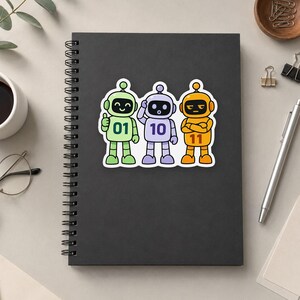 May include: A black spiral-bound notebook featuring a sticker of three cartoon robots. The green robot displays 01, the purple robot displays 10, and the orange robot displays 11. Each robot has a distinct facial expression and posture.