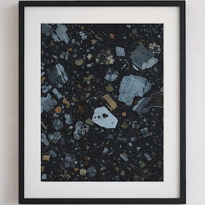 May include: A framed print featuring a detailed, abstract pattern. The artwork displays a dark, textured background with scattered fragments in shades of gray, blue, and yellow. The print is encased in a black frame with a white mat.