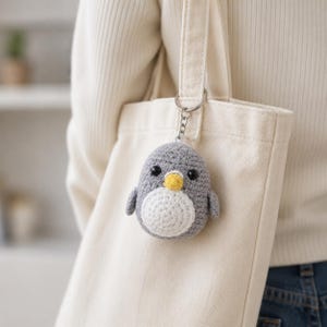May include: A small, round, crocheted penguin keychain featuring a grey body, a white belly, a yellow beak, and two black bead eyes. The plush penguin is attached to a silver-toned metal split ring and chain, hanging from a cream-colored canvas tote bag.