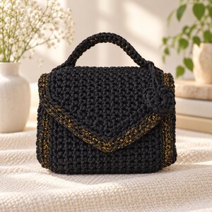 Puede incluir: Un bolso de ganchillo negro con un asa superior estructurada y un cierre de solapa triangular. El bolso est&aacute; acentuado con hilo dorado met&aacute;lico tejido a lo largo de los bordes e incluye un peque&ntilde;o dije de ganchillo circular en el asa lateral.
