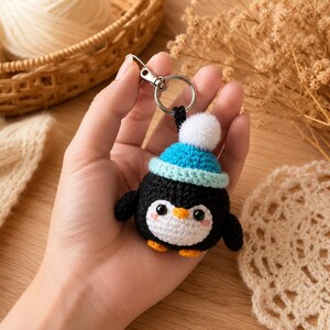 May include: A crocheted penguin keychain featuring a black body, a white belly, and orange feet. The penguin wears a bright blue crocheted hat topped with a white pom-pom. The figure has two black eyes and small pink blush marks on its cheeks, attached to a silver metal keyring.