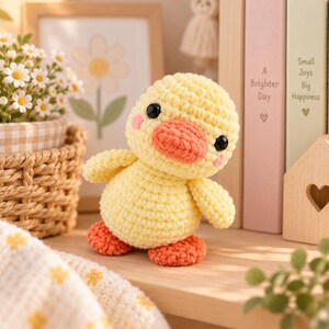 May include: A small, handmade crochet duckling toy with a round yellow body, a peach-colored beak, and two black bead eyes. The duckling has small pink blush marks on its cheeks and two orange feet. It is crafted from soft yarn with visible stitches.