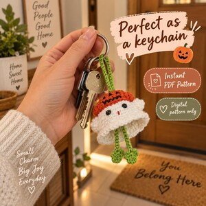 May include: A crocheted ghost keychain featuring a white body, a small orange pumpkin hat with black facial features, and two green dangling legs with clover-shaped feet. The ghost is attached to a silver metal keyring with two metal keys.