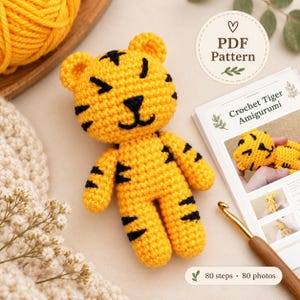 May include: A yellow and black crocheted tiger amigurumi toy with stitched facial features and stripes, accompanied by a printed PDF pattern document titled Crochet Tiger Amigurumi featuring 80 steps and 80 photos, and a wooden crochet hook.