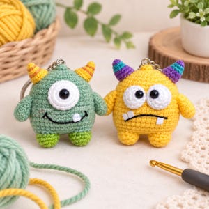 May include: Two crocheted monster keychains. The left monster is sage green with one large white eye, yellow striped horns, and a single white tooth. The right monster is yellow with two white eyes, purple and teal striped horns, and two white teeth.