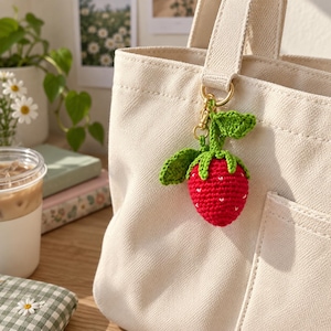May include: A cream-colored canvas tote bag featuring a crocheted strawberry keychain attached to the handle. The strawberry is bright red with small white embroidered details, topped with green crocheted leaves and a gold-toned metal clasp.