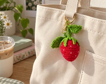 May include: A cream-colored canvas tote bag featuring a crocheted strawberry keychain attached to the handle. The strawberry is bright red with small white embroidered details, topped with green crocheted leaves and a gold-toned metal clasp.