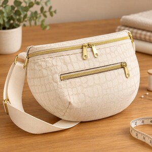 May include: A cream-colored crescent-shaped crossbody bag featuring a textured crocodile-embossed pattern, gold-tone metal zippers, and a wide adjustable fabric strap. A white measuring tape is visible in the corner of the frame.