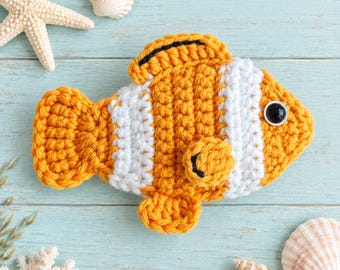 May include: A crocheted clownfish toy featuring vibrant orange and white vertical stripes, a black-rimmed dorsal fin, and a single black plastic eye. The fish is crafted with a textured yarn stitch pattern, showcasing a rounded body, a tail fin, and a small side fin.